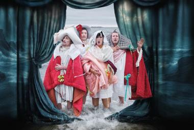 Kuvassa ovat tummien verhojen keskellä neljä näyttelijää lattiavaahdossa seisomassa. Heillä on kylpytakkimaisia valkoisia, punaisia ja vaalean punaisia täkkejä pitkien paitojen päällä. 