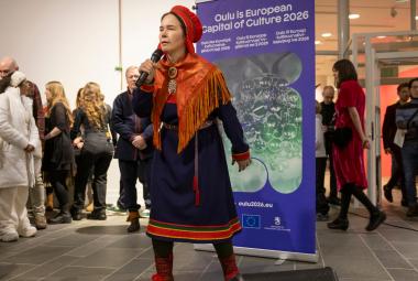 Kuvassa on Anna Morottaja keskellä kuvaa esiintymässä Lapin puvussa, joka on tumman sininen ja siihen kuuluu puna-oranssi -kuvioinen huivi ja päähine.  Taustalla näkyy Oulu2026 roll-up ja vasemmalla yleisöä.