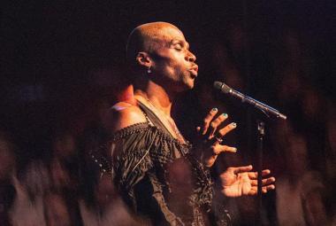 Kuvassa on Jani Toivola puolivartalokuvassa sivuttain näyttämöllä esiintymässä mikrofoni edessään. Hänellä on tumman ruskea hihaton asu päällään. Kuvan tausta on tumma.