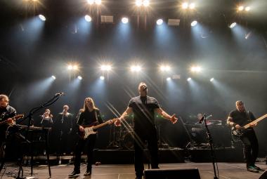 Kuvassa on neljä bändin jäsentä ja kaksi taustalaulajaa.  Lava on tumma ja taustalla näkyy valorivejä. 