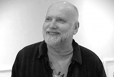 Kuvassa on Arvo Siikamäki ja hän on puolivartalokuvassa. Hänellä on tumma pusero ja vaaleahko t-paita päällään.  Hänellä on vaaleaa partaa ja päälaki on hiukseton. Kuva on musta-valkoinen.