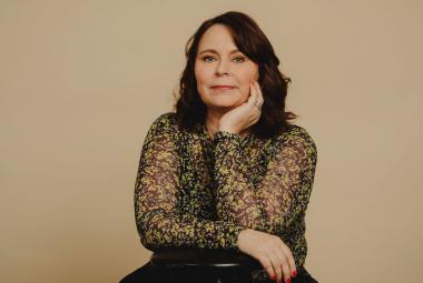 Kuvassa on Saila Susiluoto ja hän istuu sekä nojaa tuolin selkämykseen. Hänellä on ruskean mustan kirjava puseoro päällään.  Kuvan tausta on ruskehtavan harmaa. 