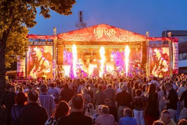 Kuvassa on takana on oranssin punainen teltta, jossa on keskellä pyrotehosteita ja vasemmalla sekä oikealla näkyy kitaristi soittamassa.  Etualalla näkyy yleisöä selin. Taustalla on kesäyön sinertävää taivasta.