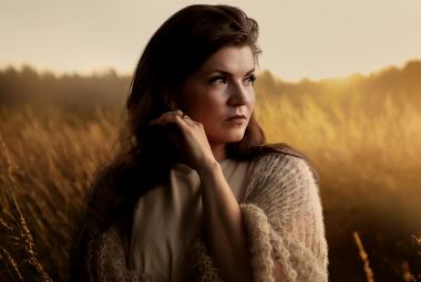 Kuvassa on Suvi Teräsniska puolivartalokuvassa ja hän on keskellä kuvaa.  Katse on oikealle. Taustalla on iltavalaistuksessa olevaa viljapeltoa.