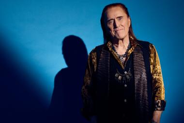 Kuvassa on Tommi Läntinen puolivartalokuvassa seisomassa. Hänellä on ruskea pusero ja musta liivi päällään. Taustalla näkyy hänen varjonsa. Vasemmalla ja alhaalla on tummennosta ja muu osa taustaa on sininen.