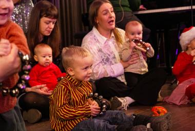 Kuvassa on äitijä ja vauvoja sekä pieniä lapsia istumassa lattialla.  He katsovat esitystä.