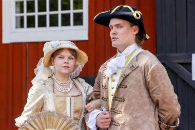 Kuvassa on vasemmalla nuori nainen vaalean ruskeassa vanhanaikaisessa mekossa ja lierihatussa. Asussa ja hatussa on valkoisia röyhelöitä ja brodyyrejä. Oikealla on nuori mies, jolla on musta aatelisen suikkahattu päässään sekä koristeellinen vaalean ruskea takki päällään ja valkoinen röyhelöpaita. Taustalla on punaisen puurakennuksen seinää ja valkoinen, pieniruutuinen ikkuna.