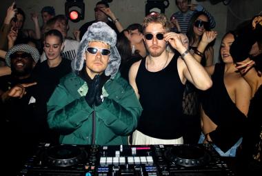 Kuvassa on keskellä vasemmalla VilleGalle vihreässä toppatakissa ja harmaassa karvalakissa sekä aurinkolaseissa. Oikealla hänen vieressään on mustassa hihattomassa t-paidassa sekä aurinkolaseissa. Taukana on nuorta yleisöä. Edessä on musiikin ohjauspöytä. 