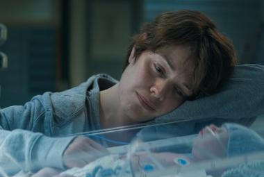 Kuvassa on Oona Airola, joka katselee keskoskaapissa nukkuvaa vauvaa. Hänellä on vaalean sininen pusero päällään. 