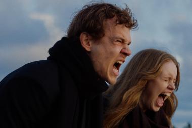 Kuvassa ovat nuori mies ja nainen puolivartalokuvassa ulkotakeissa. He molemmat ovat sivuttain ja huutavat ulkona. Taustalla on puolipilvistä taivasta.