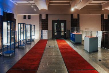 Kuvassa on Helsingin päärautatieaseman näyttelytila, jossa on isossa tilassa molempien seinien luona lasiset vitriinihyllyt. Seinät ja lattia ovat vaalean ruskeat ja keskellä on kaksi pitkää punaista mattoa.