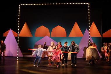 Kuvassa on tummalla näyttämöllä takana punaisia kartiomaisia telttoja. alemana on näytteljöitä tanssimassa edessä värikkäissä vaatteissa ja taaempana seisoo harmassa puvuissa olevia näyttelijöitä. Vasemmalla on kartiomainen lila teltta.