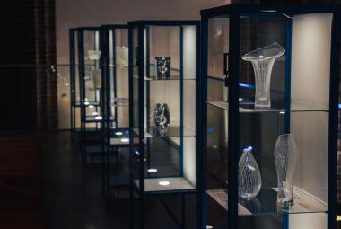 Kuvassa on tummassa tilassa keskellä läpinäkyvästä lasista hyllykkö, jossa on Tapio Virkkalan lasitöitä.
