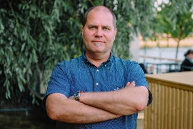 Kuvassa on keskellä Stuba Nikula seisomassa puolivartalokuvassa.  Hänellä on sininen, lyhythihainen  paita päällään ja hänellä on kädet toistensa päällä.  Taustalla on alaspäin laskeutuvia lehtipuun oksia.