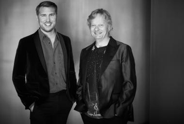 Kuvassa on seisomassa vasemmalla aarne Pelkonen mustassa takissa ja harmaassa paidassa kädet taskussa. Oikealla on Iiro Rantala mustassa kiiltävässä takissa. Tausta on tumma ja siinä on taustavaloa. Kuva on musta-valkoinen.