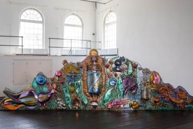 Kuvassa on isossa valkoisessa tilassa, jossa on vasemmalla ylhäällä isoja, yläosasta pyöreitä ja pikkuryytuisia ikkunoita. Tummalla lattialla on kiiltävävärisiä, erityisesti turkoosia, ruskeaa ja lilaa ison krokotiilimaisen hahmon päällä.  Hahmo nousee ylöspäin ja laskeutuu alas.  Päällä on keraamisia kuvioita ja mm. lintuja.