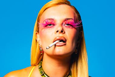 Kuvassa on puolivartalokuvassa Erika Vikman ja hänellä on pitkät vaaleat hiukset, punaiseksi meikatut silmänympärykset ja tupakka hampaissaan. Tausta on turkoosi. 