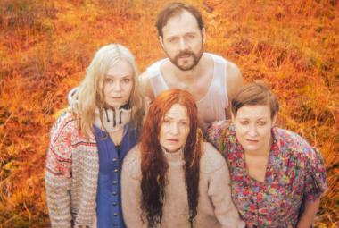 Kuvassa on ylhäältä päin kuvattuna edessä kolme naisnäyttelijää ja takana yksi mies. He ovat tiiviinä ryhmänä. Tausta on oranssi.