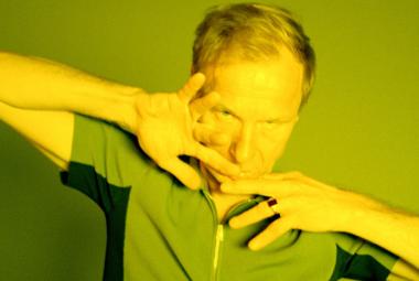 Kuvassa on Ismo Alanko puolivartalokuvassa ja hänellä on kämmenet viistosti kasvojen päällä. Kuvan sävy on vihreä.