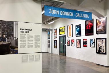 Kuvassa on julistegalleria, joussa on etuseinällä kuvaa ja tekstiosuus. Käytävän toisella puolella on julisteita ja ylläpuolella sininen kyltti, jossa on valkoisin kirjaimin Jörn Donner -galleria.