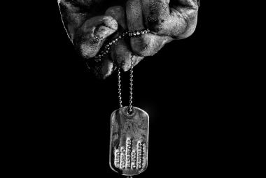 Kuvassa on ylhäältä päin tuleva käsi, josta roikkuu pieni kultalaatta. Kuva on musta-valkoinen ja tausta on musta.