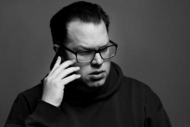 Kuvassa on Jonas Olson puolivartalokuvassa. Hänellä on oikeassa kädessään kännykkä korvallaan. Hänellä on tummasankaiset silmälasit ja musta poolokauluspusero päällään. Tausta on harmaa.