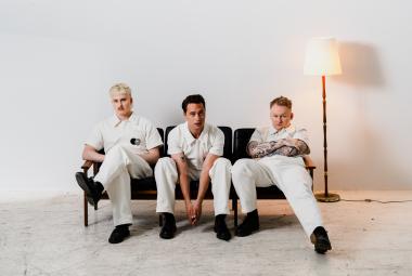 Kuvassa ovat Kuumaa-yhtyeen kolme jäsentä istumassa sihvalla. Heillä kaikilla on valkoiset vaatteet. Oikealla on jalkalamppu. Kuvan tausta on valkoinen ja lattia vaalea.