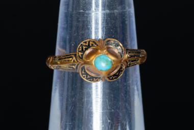 Kuvassa on tummahko koristeltu kultasormus, jossa on keskellä turkoosi kivi ja sen ympärillä leveitä koristeita. Sormus on läpinäkyvän putkilon ympärillä.  Tausta on tumma.