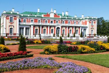 Kuvassa on Kadriorgin taidemuseon rakennus, joka on puna-valkoinen ja siinä on korkeat koristeelliset ikkunat ja valkoinen koko julkisivun yläparveke. Edessä on värikkäitä kukkaistutuksia.