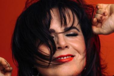 Kuvassa on Kaija Koo kasvokuvassa. Hänellä on tummat hiukset osittain kasvoilla.  Tausta ruskean punainen.