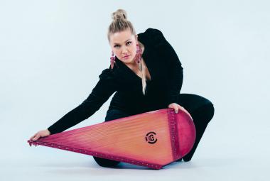 Kuvassa on Maija Kauhanen mustassa asussa kumartuneena punaisen vasemmalle suipoksi muotoillun kanteleen takana ja hän pitää oikealla kädellään kanteleen päästä kiinni. Tausta on vaalean sininen.  