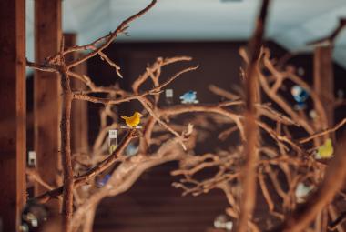 Kuvassa on ruskeissa pystyrungoissa ruskeita puun oksia, joissa on pieniä eri värisiä lasilintuja.