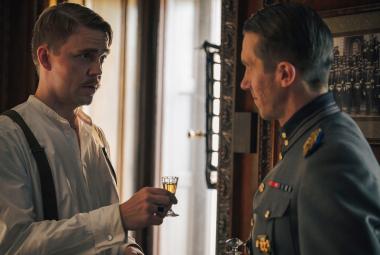 Kuvassa on vasemmalla Aku Sipola valkoisessa paidassa kohottamassa lasia sotilaspuvussa olevalla Jussi Vataselle, joka on osittain selin. Taustalla on iso ikkuna ja kuva sävy on tummahko. 