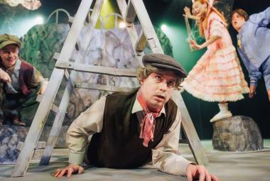 Kuvassa on nuori mies lippalakissa ja tummassa liivissä sekä vaaleassa paidassa. Hän ryömii harmaiden kolmiotikkaiden ali. Oikealla istuu poika kyykyssä kiven päällä ja hänellä on samanlaiset vaatteet. Oikealla näkyy tytön vaalean oranssi rimpsuhame. Hänen oikealla puolellaan on pikku poika vaalean sinisessä housupuvussa.