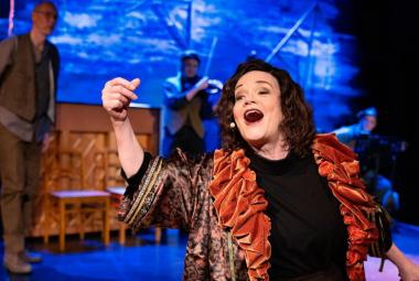 Kuvassa on Lise Holmberg keskellä laulamassa mustassa mekossä ja ruskehtavassa rimpsujakussa. Taustalla on  vasemmalla mies ruskehtavissa vaatteissa ja takana on soittaja.