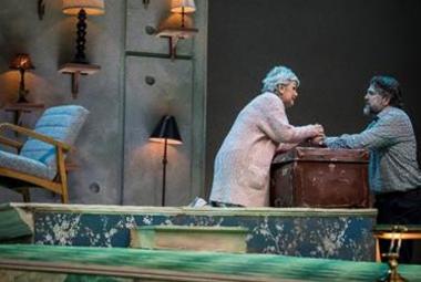 Kuvassa on huone ja vasemmalla nojatuoli sekä seinällä lamppuja. Oikealla on ruskean tavara-arkun ääressä seisomassa kumarassa naisen ja toisella puolella on mies.