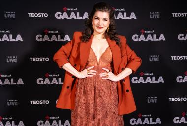 Kuvassa on Suvi Teräsniska tumman oranssin ruskeassa mekossa ja jakussa.  Hänellä on kädet lanteillaan. Taustalla on tummapohjaisella seinällä ja siinä lukee moneen kertaan Gaala ja Teosto.