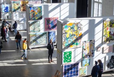 Kuvassa on isossa tilassa kolme isoa teosjalustaa ja niissä kaikissa useita tauluja. Jalustojen keskellä on ihmisiä katsomassa teoksia ja valoa heijastuu ikkunoista vinottain teosten päälle.