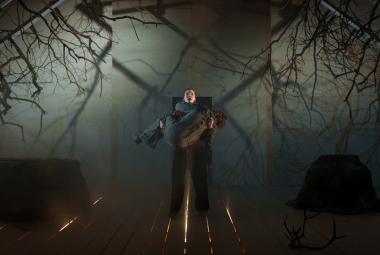 Kuvassa on keskellä Tommi Eronen kantamassa Tuuli Pajua.  Kuvan sävy on tummala ylhäällä on oksia roikkumassa puupalkeista molemmin puolin ja taustalla on sumua. 