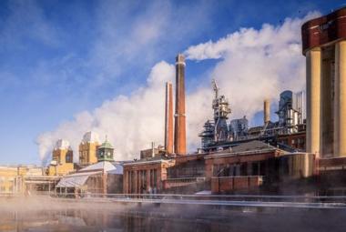 Kuvassa on edessä vettä ja takana punatiilisiä tehdasrakennuksia, joissa on torneja ja oikealla korkeat savupiiput, joista tulee savua. Tautalla on sinistä taivasta.