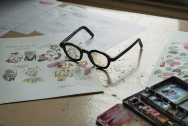 Kuvassa on pilapiirroskuvia pöydällä valkoisella paperilla ja niiden päällä silmälasit. Oikeassa reunassa on värejä.