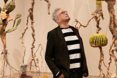 Kuvassa on keskellä Roland Persson seisomassa puolivartalokuvassa. Hänellä on musta takki ja musta-valkoinen paksuraidallinen pusero päällään. Hän katsoo ylöspäin. Taustalla on roikkuvia juurista tehtyjä taideteoksia ja seinät ovat vaalean ruskeat.