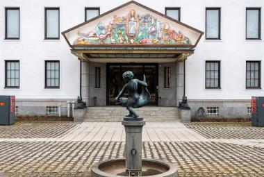 Kuvassa on valkoinen rakennus jossa on kapeita ikkunoita kahdessa kerroksessa. Siinä on portaikko ja isot ovet ja niiden päällä iso kolmio. Siinä on reliefimäisesti palstellivärisiä hahmoja ja esineitä.