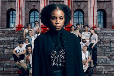 Kuvassa on keskellä nuori nainen mustassa kaavussa, jossa on hopeinen perhonen edessä. Hänellä on mustat, kiharat hiukset. Taustalla on valoisissa puseroissa istumassa harmailla portailla joukko nuoria. 