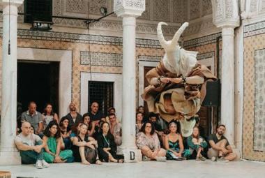 Kuvassa on joukko ihmisiä istumassa rakennuksen valkoisella lattialla ryhmänä ja edessä on kolme valkoista, itämaista pylvästä. taustalla näkyy seinää, jossa on valkoinen, avoin oviaukko ja vieressä kaksi pienempää aukkoa. Seinässä on keltakuvioista mosaiikkia ja sinikuvioista ornamenttia. 