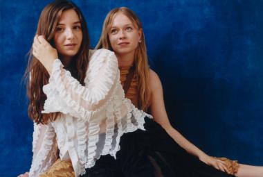 Kuvassa ovat Emma ja Matilda istumassa lattialla vinoasennossa. Emma on vasemmalla edessä ja hänellä on valkoinen hapsupusero. Hänen takanaan on Matilda, joka on hihattomassa puserossa.