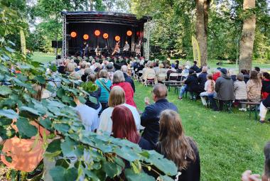 Kuvassa on puiston vihreitä puita ja oksia. Taustalla on esiintymislava ja siinä orkesteri soittamassa. Lavan edestä taakse päin on yleisöä istumassa tuoleilla.