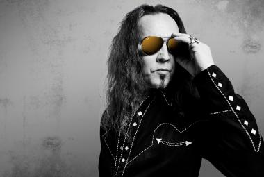 Kuvassa on Jukka ahola puolivartalokuvassa ja hänellä on musta takki päällään. Siinä on nappeja hihassa ja Ahola pitää vasemmalla kädellään aurinkolaseista kiinni. Kuva on musta-valkoinen tausta on harmaa.