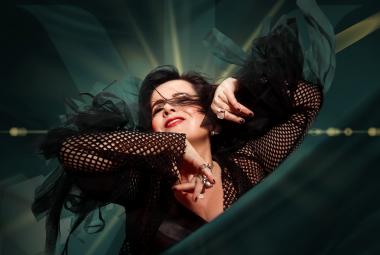 Kuvassa on Kaija Koo puolivartalokuvassa ja hänellä on kädet kohotettuina vauhdikkaasti. Hänellä on musta puku ja hihat ovat verkkomaiset. Tausta hihreän tumma ja valoa tulee pään yläpuolelta.