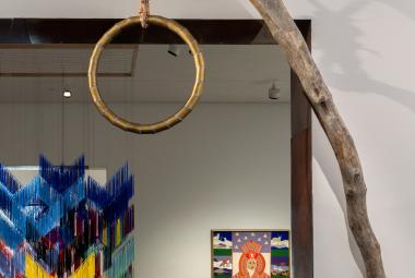 Kuvassa on etualalla kapeaa puunrunkoa ja siitä roikkuu puinen, pyöreä hirttosilmukka. Taustalla näkyy roikkuva tekstiilityö, jossa on Lapin värejä ja näyttelysalia.